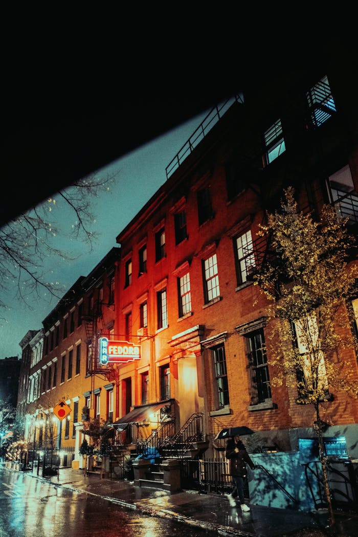 Illuminated city street at night with buildings and glowing signs on a rainy evening.
